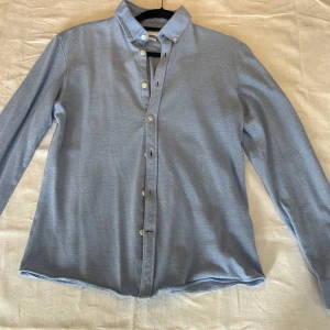 Ljusblå skjorta Lindbergh White  - Snygg ljusblå skjorta från Lindbergh White i slim fit-modell. Skjortan har klassisk krage med knappar och långa ärmar. Storlek M. Lite tjockare i materialet så passar bra året om. Perfekt för dig som gillar stilrena och enkla plagg. Materialet känns mjukt och andas bra.