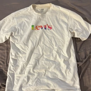 Vit Levi's t-shirt med färgglad logga - Vit t-shirt från Levi's i relaxed passform. Framsidan har en stor, färgglad Levi's-logga i regnbågens färger. Klassisk rund hals och korta ärmar. Tillverkad i mjuk bomull, perfekt för chill dagar och streetstyle.