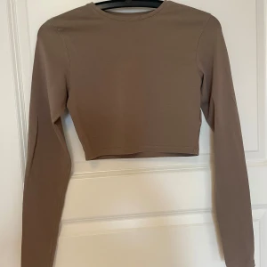 Brun croppad långärmad topp - Snygg croppad topp i en mjuk brun färg med lång ärm och rund hals. Enkel och stilren design utan mönster eller detaljer, perfekt att matcha med jeans eller kjol. Tillverkad i stretchig bomull som sitter skönt mot kroppen.