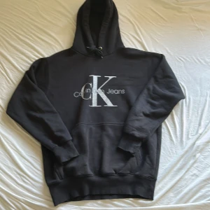 Svart Calvin Klein hoodie med logga - Svart hoodie från Calvin Klein Jeans med stor CK-logga i vitt på bröstet. Tröjan har huva med snörning, ribbade muddar och klassisk känguruficka framtill. Perfekt för chill dagar och streetwear-stil. Materialet är mjuk bomull och polyesterblandning. Storlek 170cm nypris 900