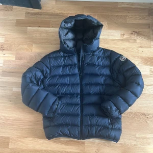 Mörkblå Colmar dunjacka med huva - Snygg mörkblå dunjacka från Colmar med huva och quiltad design. Jackan har dragkedja framtill, två fickor med dragkedja och en rund Colmar-logga på ärmen. Perfekt för kalla dagar och riktigt skön att bära.