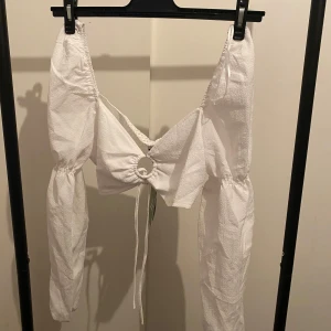 Vit omlottopp från H&M Divided - Trendig vit omlottopp från H&M Divided med långa puffärmar och snygg ringdetalj framtill. Toppen har knytning och smockad rygg för extra komfort. Tillverkad av 87% återvunnen polyester, perfekt för dig som gillar en cool och modern stil.