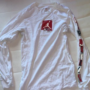 Vit långärmad Jordan tröja - Snygg vit långärmad tröja från Jordan med rött Air Jordan-logga på bröstet och tryck längs ena ärmen. Skön passform och tillverkad i mjuk bomull, perfekt för en sportig och avslappnad stil.