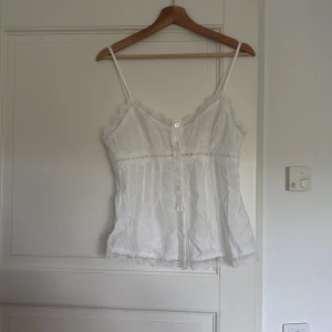 Vit spetsdetaljerad linnetopp - Supersöt vit linnetopp med tunna axelband, knappar framtill och fina spetsdetaljer längs kanterna och över bysten. Lätt och luftig med romantisk känsla, perfekt för varma dagar eller lager på lager. Ger en fräsch och feminin vibe.