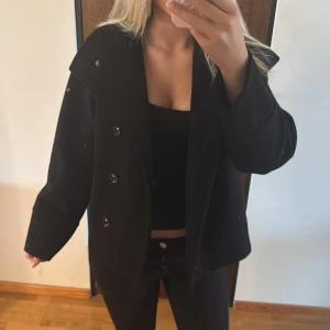 Svart skepparkavaj från Gina Tricot - Stilren svart skepparkavaj från Gina Tricot med bred krage och dubbelknäppning framtill. Jackan har en rak passform och är tillverkad i ett mjukt material som känns skönt. Storlek 146/152!