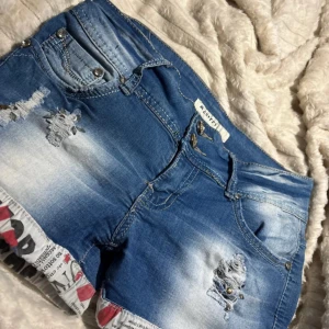 Blå jeansshorts från Mcherry - Snygga blå jeansshorts från Mcherry med slitna detaljer och coola tryckta kanter i tidningsstil. Shortsen har dubbla knappar fram, fem fickor och kontrastsömmar. Passformen är normal och materialet är mestadels bomull med lite elastan för extra stretch.