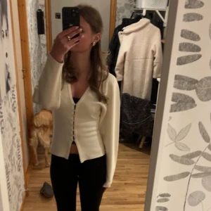 Beige kofta med knappar och v-ringning - Snygg beige kofta med v-ringad hals och dekorativa knappar framtill. Modellen är figurnära med långa ärmar och mjukt stickat material som känns skönt mot huden. Perfekt att styla med jeans eller kjol för en clean look.