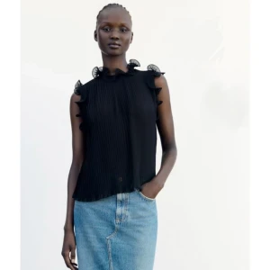 Svart plisserad blus med volang - Säljer en svart blus med plisserat tyg och volangdetaljer vid axlar och halsringning. Blusen är ärmlös och har en lätt transparent känsla, vilket ger en cool och luftig vibe. Perfekt att styla med jeans eller kjol för en trendig look.