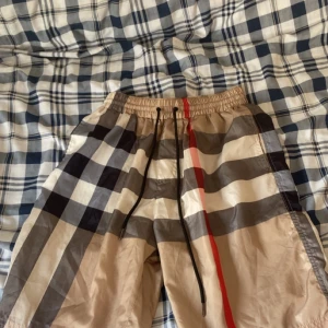 Burberry rutig shorts med snörning - Snygga Burberry shorts i klassiskt rutigt mönster med beige, svart, vit och röd färg. De har elastisk midja med svart snörning och är tillverkade i 100% bomull. Perfekt för dig som vill sticka ut med designerplagg och cool stil.
