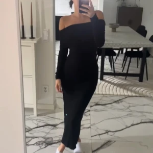 Svart offshoulder långklänning - Snygg svart långklänning med offshoulder och lång ärm. Klänningen har en tight passform och en liten slits baktill för extra rörelsefrihet. Perfekt för dig som gillar stilrena och eleganta plagg med en modern touch.