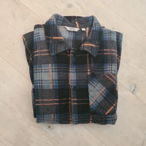 Rutig overshirt från Warp i blått - Snygg rutig overshirt från Warp i blå, svart och orange. Skjortan har klassisk krage, bröstficka och knäppning framtill. Perfekt att slänga över en t-shirt för en chill look. Materialet är mjukt och känns skönt mot huden.