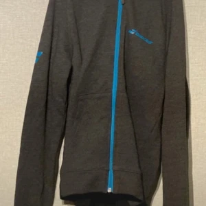 Svart hoodiejacka från Babolar S - Svart hoodiejacka från Babolar med blå dragkedja och logga på bröstet och ärmen. Jackan har huva, långa ärmar och är tillverkad i en mjuk bomullsblandning med polyester. Perfekt för dig som gillar sportig stil och vill ha något bekvämt till träning eller chill.