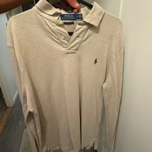 Beige långärmad pikétröja Polo Ralph Lauren - Klassisk beige långärmad pikétröja från Polo Ralph Lauren med broderad logga på bröstet. Tröjan har krage, knappar vid halsen och är tillverkad i mjuk bomull. Perfekt för en stilren och avslappnad look.
