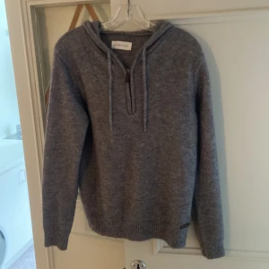 Grå stickad hoodie från Pier One - Mysig grå hoodie från Pier One med huva och snörning. Tröjan är stickad och har en half zip-dragkedja framtill. Långärmad modell med ribbade muddar vid ärmslut och nederkant. Perfekt för kyliga dagar när du vill vara både chill och snygg.