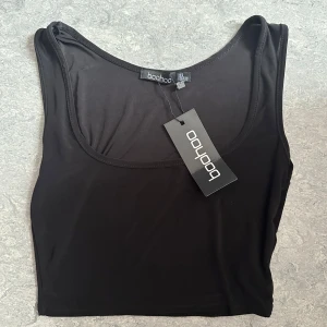 Svart croppad linnetopp från Boohoo strl Xs - En svart, croppad linnetopp från Boohoo med rund halsringning och ärmlös design. Toppen är i ett mjukt och stretchigt syntetmaterial som sitter snyggt mot kroppen. Perfekt att styla med jeans eller kjol för en clean look.