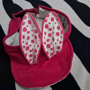 Rosa keps med kaninöron och prickar - Söt rosa keps med två stora kaninöron framtill. Öronen är vita med rosa prickar och har rosa kant. Kepsen har justerbart band bak och är tillverkad i mjuk bomull. Fint skick:)