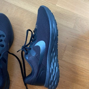 Svarta Nike sneakers med mesh - Snygga helsvarta Nike sneakers med diskret logga på sidan. Ovandelen är i ventilerande mesh och sulan är rejäl och bekväm. Perfekta för dig som gillar en clean look och vill ha ett par lätta skor till vardagen eller träningen.