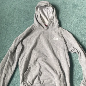 Grå hoodie från The North Face - Säljer en grå hoodie från The North Face med stor logga och texten 'Never Stop Exploring' på ryggen. Tröjan har huva, känguruficka och långa ärmar. Perfekt för chill dagar eller när du vill ha en enkel streetstyle-look.