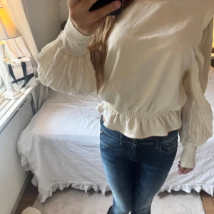 Vit blus med volang och puffärm - Supersöt vit blus med breda volangdetaljer i midjan och på de långa puffärmarna. Blusen har resår i midjan som ger en snygg form och extra volym i ärmarna. Xs-s