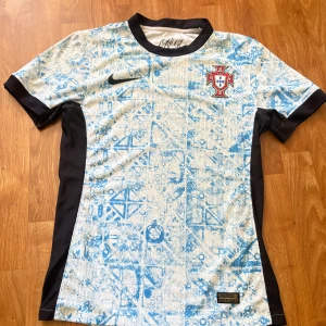 Portugal Nike fotbollströja blå/vit - Portugal landslagströja från Nike med unikt blått mönster på vit bas, svarta detaljer på sidor och ärmslut. Broderad landslagssköld på bröstet och Nike-logga. Tillverkad i lätt, ventilerande Dri-FIT-material som passar perfekt för träning eller match. 200 är sista pris.