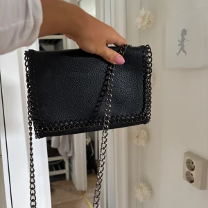 Svart axelväska med kedjerem - Snygg svart axelväska i skinnimitation med grov kedjerem och dekorativ kedjedetalj runt kanterna. Stilren och edgy design som passar perfekt till streetwear eller en cool vardagslook.