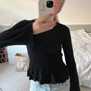 Svart smockad topp med volang - Svart långärmad topp från Gina Tricot med smockad front, volangkant nedtill och utsvängda armar. Passar även xs. Använd endast några fåtal gånger!