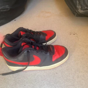 Nike Dunk Low svart/röd sneakers - Snygga Nike Dunk Low sneakers i svart och rött skinn med klassisk röd swoosh och svart snörning. Låg modell med vit sula och perforerad tåbox för extra stilpoäng. Perfekta för dig som vill sticka ut med en ikonisk streetwear-look.