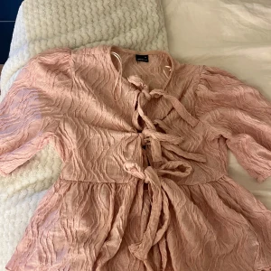 Ljusrosa blus med knyt och struktur - Supersöt ljusrosa blus från Gina Tricot med crinklad struktur och tre knyt framtill. Modellen har korta puffärmar och en lätt utsvängd passform som ger en romantisk vibe. Perfekt att styla med jeans eller kjol för en trendig look.💞 Har bara använt en gång!