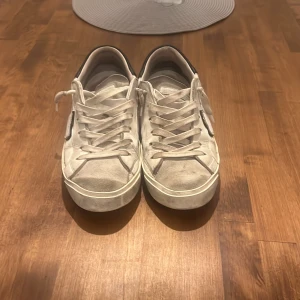 Vita Philippe Model sneakers i skinn - Snygga vita sneakers från Philippe Model med svart detalj på hälen och klassisk snörning. Skorna har en rund tå, platt sula och är tillverkade i skinn. Ikonisk sköld-logga på sidan och diskret branding på baksidan. Perfekta för en clean och stilren look.