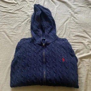 Mörkblå stickad hoodie från Polo Ralph Lauren - Säljer en mörkblå stickad hoodie från Polo Ralph Lauren med dragkedja och huva. Tröjan har ett klassiskt kabelstickat mönster, ribbade muddar och fickor framtill. Den ikoniska röda Polo-loggan är broderad på bröstet. Perfekt för dig som gillar chill och stilren look.