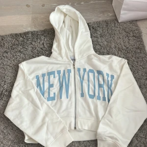 Vit hoodie med blått New York-tryck - Säljer en vit croppad hoodie från Zara  med stort ljusblått New York-tryck på framsidan. Tröjan har huva, dragkedja och långa ärmar med ribbade muddar. Perfekt för dig som gillar streetstyle och vill ha en chill look.
