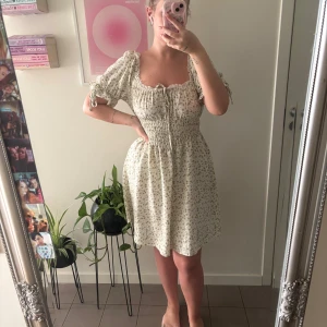 Blommig kort klänning med spets - Supersöt kort klänning i ljus beige med små gröna blommor, puffärmar med knyt och smock i midjan. Urringningen har fina spetsdetaljer och snörning framtill. Perfekt för sommarens alla dagar, mjukt och lätt material. Storlek L men väldigt strechig 