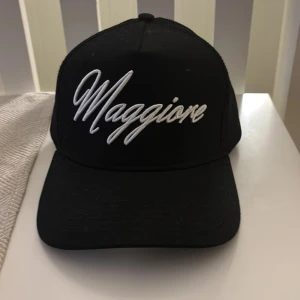 Svart Maggiore keps  - Svart keps från Maggiore med vit broderad logga framtill. Klassisk trucker-modell med böjd skärm och justerbar snapback. Baksidan är i mesh för extra ventilation och insidan har svarta band med Maggiore-tryck.