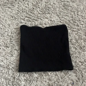 Svart ribbad bandeau tubtopp - En svart bandeau tubtopp med ribbad struktur. Toppen är axelbandslös och har en enkel, stilren design som är lätt att matcha med olika outfits. Perfekt för dig som gillar minimalistisk stil och vill ha en topp som sitter tight och snyggt.