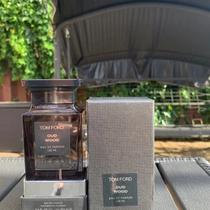 Tom Ford Oud Wood EdP 100ml - Tom Ford Oud Wood Eau de Parfum 100 ml med lyxig mörkbrun glasflaska och elegant svart etikett. Flaskan har en fyrkantig, stilren design och levereras med originalkartong i grått mönstrat material. En ikonisk doft med exklusiv känsla.