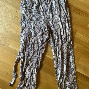 Ormmönstrade byxor med knytband - Säljer ett par flowiga byxor med ormmönster i beige och bruna toner. Byxorna har elastisk midja och knytband i midjan för extra detalj. Perfekta för dig som vill sticka ut med ett djurmönster och gillar en avslappnad vibe.