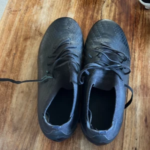 Svarta fotbollsskor från Adidas - Säljer ett par svarta fotbollsskor från Adidas med coola mönstrade dobbar undertill. Skorna har snörning och är tillverkade i syntetmaterial. Perfekta för dig som vill ha bra grepp på planen och en stilren look.
