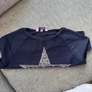 Mörkblå tröja med paljettstjärna -Stockholm stil - Snygg mörkblå tröja från SVO med en stor stjärna i silverfärgade paljetter på framsidan. Tröjan har rund hals och korta ärmar, perfekt för dig som gillar lite extra bling i vardagen.  