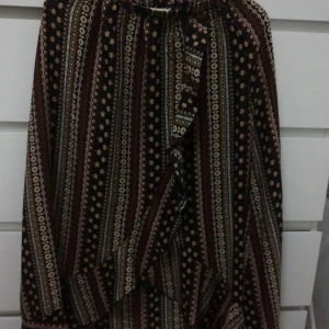 Mönstrad långkjol med volang - Säljer en boho-inspirerad långkjol med vertikala ränder i svart, beige och vinrött. Kjolen har ett coolt mönster med små blommor och detaljer, samt en volang framtill som ger extra flow. Perfekt för dig som gillar unika prints och avslappnad stil.