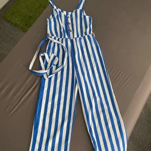 Randig blåvit byxdress med knappar - Snygg byxdress med blå och vita vertikala ränder, knappar framtill och tillhörande bälte i midjan. Vida ben och smala axelband ger en chill och somrig vibe. Perfekt för dig som vill sticka ut med färg och mönster.