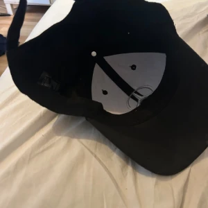 Svart Mercedes keps med broderad logga - Snygg svart keps med klassisk böjd skärm och justerbart spänne bak. Framtill sitter en broderad Mercedes-logga i silver. Kepsen är tillverkad i bomull och har ventilerande hål upptill för extra komfort. Perfekt för dig som gillar stilrena accessoarer och bilar.