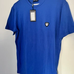 Blå EA7 t-shirt från Emporio Armani - Snygg blå t-shirt från EA7 Emporio Armani med rund hals och korta ärmar. T-shirten har en stilren logotyp på bröstet och är gjord i mjuk bomull som känns skön mot huden. Perfekt för dig som gillar klassisk design med en modern touch.
