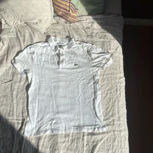 Vit Lacoste pikétröja slim fit M - Vit pikétröja från Lacoste i slim fit-modell med klassisk krage och knappar framtill. Ikoniska gröna krokodilloggan broderad på bröstet. Tillverkad i bomull, perfekt för en clean och sportig look. Lite använde men inga fläckar.
