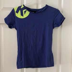 Blå t-shirt Kempa  - Blå t-shirt från Kempa med neonfärgat tryck på axeln. Modellen är klassisk med rund hals och korta ärmar. Materialet är mjuk bomull som känns skönt mot huden. Perfekt för dig som gillar sportig och enkel stil.