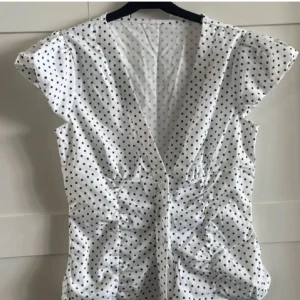 Vit prickig blus med v-ringning - Supersöt vit blus med svarta prickar och djup v-ringning. Korta puffärmar och knappar framtill ger en feminin vibe. Figurnära passform med rynkade detaljer i midjan. Perfekt till jeans eller kjol för en trendig look.