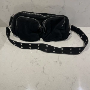 Svart axelväska med nitar och dragkedjor - Köp via instabox!! Cool svart axelväska i mocka med bred axelrem dekorerad med silvriga nitar. Väskan har flera fack med grova dragkedjor i silver och en mjuk, lite puffig form. Perfekt för dig som vill ha en edgy accessoar till din outfit.