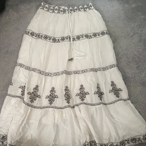 Vit långkjol med broderade detaljer - Boho-inspirerad vit långkjol med svarta broderade mönster och snörning i midjan. Kjolen har flera volanger och ett unikt mönster längs kanterna och nedtill. Perfekt för dig som gillar en avslappnad och stilren look. Nypris ca 600kr