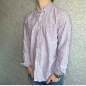 Ljuslila skjorta från Polo Ralph Lauren - Säljer en stilren ljuslila skjorta från Polo Ralph Lauren med klassisk button-down krage och broderad logga på bröstet. Skjortan är långärmad, har normal passform och är tillverkad i mjuk bomull. Perfekt för dig som gillar en clean och tidlös look.