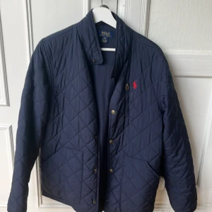 Mörkblå quiltad jacka Polo Ralph Lauren - Snygg mörkblå quiltad jacka från Polo Ralph Lauren med klassisk röd logga på bröstet. Jackan har ståkrage, tryckknappar samt två fickor framtill. Perfekt för höst och vår när du vill ha en stilren look. Slitage på båda fickorna och en av knapparna har åkt av