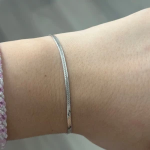 Tunt silverarmband från ANI - Snyggt och minimalistiskt armband i silverfärgad metall med platt, glänsande kedja. Armbandet har ett litet grönt hänge vid låset som ger en extra detalj. Perfekt för dig som gillar stilrena smycken med en liten twist.